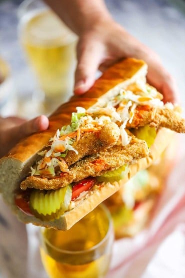 A person holding a large fried catfish po-boy that is stuffed with crispy fillet of catfish, coleslaw, hot sauce, pickles, and tomatoes.