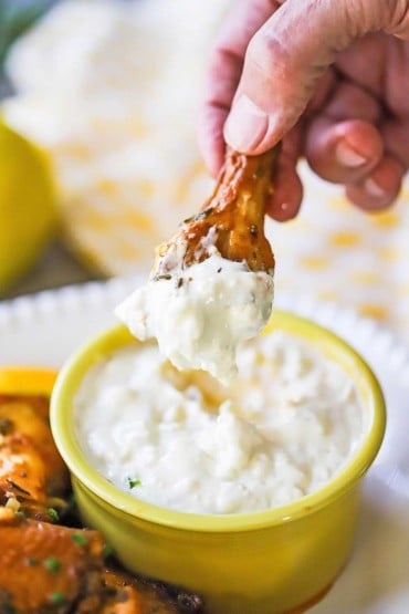 A person holding an Italian-style chicken wing that has been plunged and removed from a yellow bowl of gorgonzola dipping sauce.