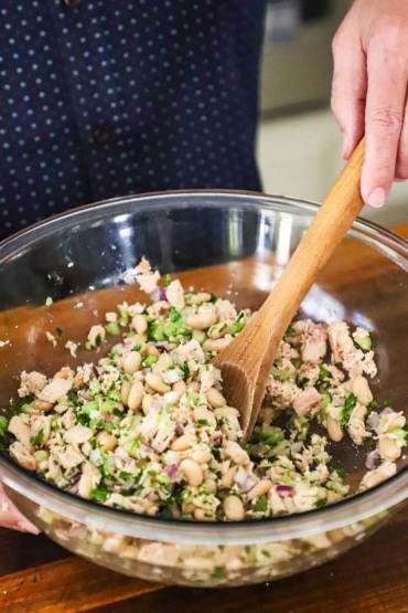 A person using a wooden spoon to mix together an albacore tuna and white bean salad in a large glass bowl.
