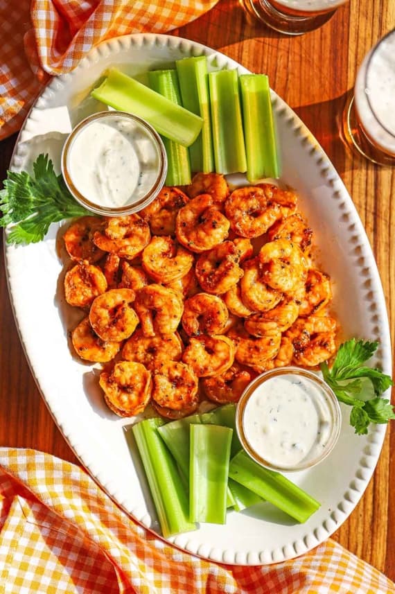An overhead view of a white oval platter filled with grilled Buffalo shrimp surrounded by strips of celery and two small jars filled with blue cheese dressing.