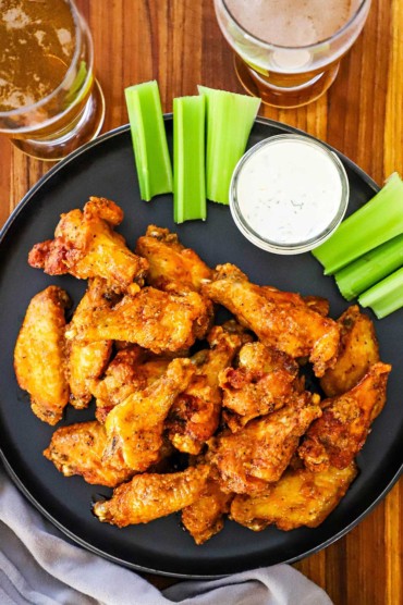 An overhead view of pile of classic Buffalo wings on a black circular plate also holding celery sticks and a small jar of blue cheese dressing.