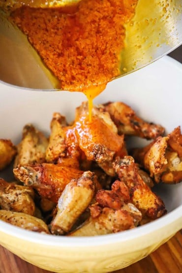 Homemade Buffalo sauce pouring from a silver saucepan into a ceramic bowl filled with fried chicken wings.