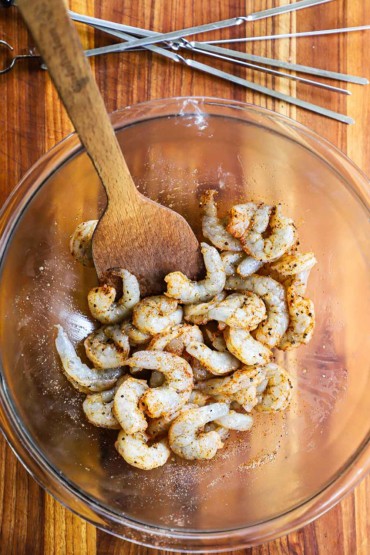 An overhead view of a glass bowl filled with uncooked shrimp that is coated with seasonings and a wooden spoon inserted on the side.