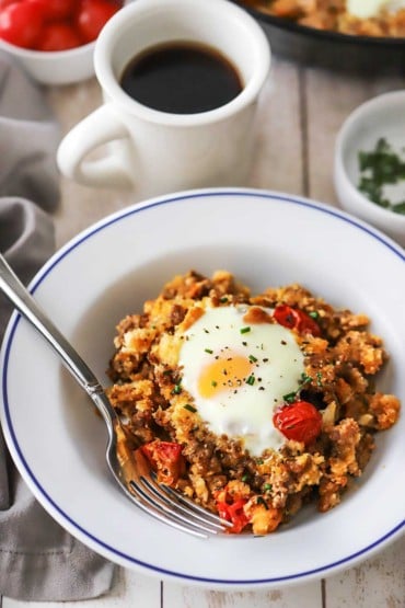 An overhead view of a white bowl filled with a helping of skillet breakfast with a mug of black coffee nearby.