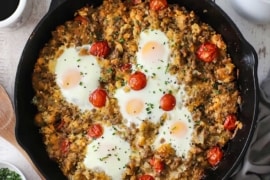 An overhead view of a skillet breakfast of cooked sausage mixed with crumbled biscuits, cherry tomatoes, and cheese all topped with five cooked eggs.