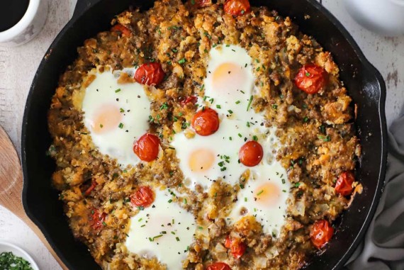 An overhead view of a skillet breakfast of cooked sausage mixed with crumbled biscuits, cherry tomatoes, and cheese all topped with five cooked eggs.
