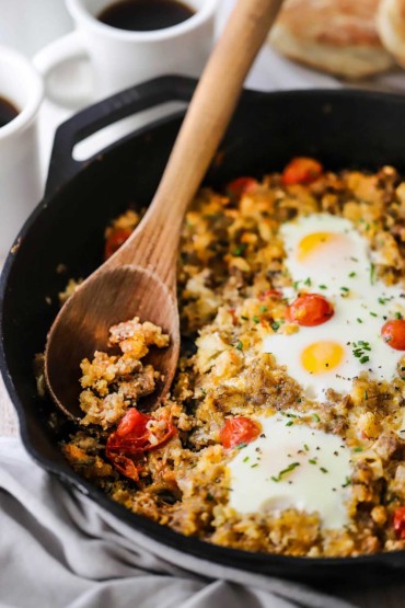 A straight-on view of a cast-iron skillet filled with a sausage hash with cooked eggs on top, and a wooden spoon inserted where some of the breakfast mixture is missing.