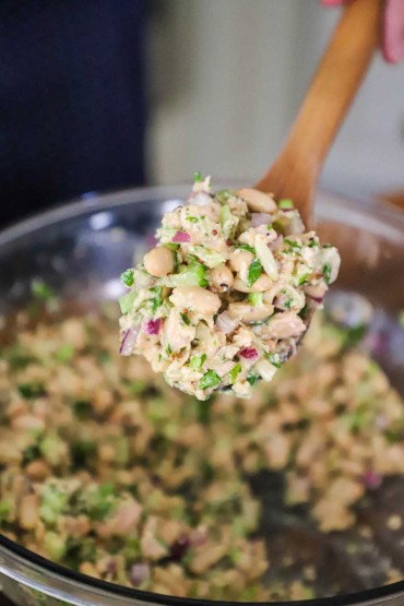 A person raising a wooden spoon filled with a tuna and white bean salad from a glass bowl of the same.