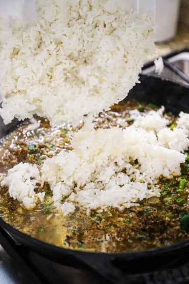 A large bowl of cooked white rice being transferred into a large skillet of a simmering sauce with cooked sausage, scallions, and herbs.