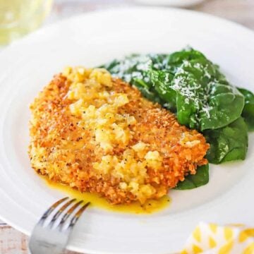 A close-up view of an almond crusted cod with orange sauce next to a small pile of a spinach salad on a white dinner plate.