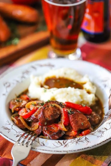 A straight-on view of an antique bowl filled with beer-braised kielbasa and pepper and mashed potatoes sitting next to a tall glass of amber beer.