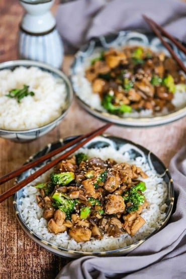 A straight-on view of a shallow bowl filled with steamed white rice topped with chicken and broccoli stir-fry with chop sticks on the side of the bowl.