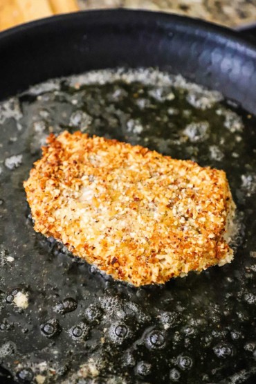 A fillet of cod that has an almond and Parmesan breading on it and is a cast-iron skillet lightly frying in olive oil.