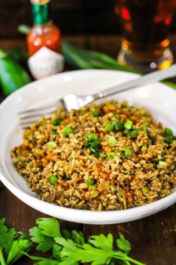 A straight-on view of a white serving bowl filled with Cajun dirty rice topped with chopped scallions and several dashes of cayenne hot sauce.