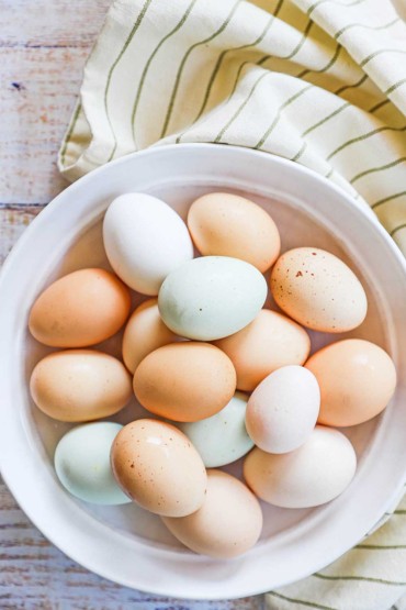 An overhead view of a white ceramic bowl filled with farm fresh eggs ranging in colors from beige, white, and a faint blue.
