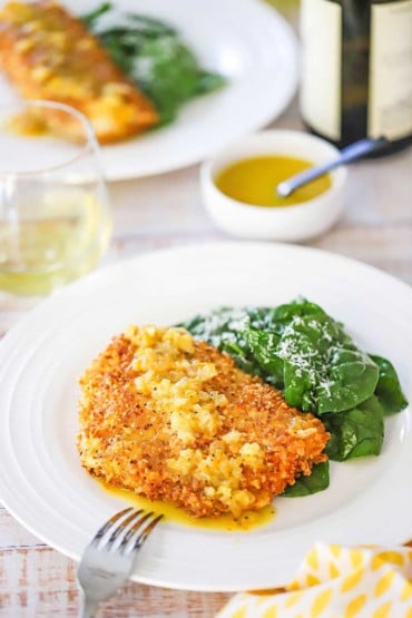 A straight-on view of a white plate with a serving of almond crusted cod sitting next to a spinach salad topped with grated parmesan.