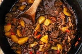 An overhead view of a black cast-iron pot filled with Hungarian goulash with chunks of beef, yellow potatoes, red bell peppers, and a brown sauce.