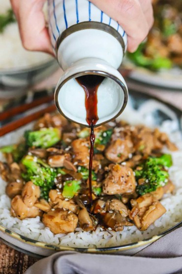 A person pouring soy sauce from a small ceramic jug onto a bowl filled with chicken and broccoli stir-fry over steamed white rice.