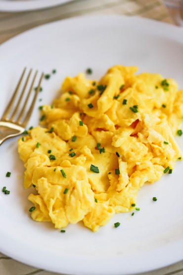 A close up view of the fluffiest scrambled eggs on a white plate and are topped with finely snipped chives.