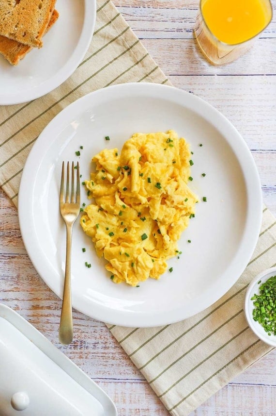 An overhead view of fluffiest scrambled eggs on a white plate topped with finely snipped chives and a gold fork on the plate, too.