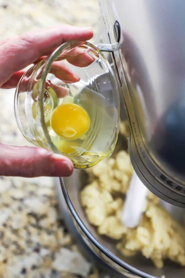 A person transferring a whole egg from a small glass bowl into the bowl of a stand mixer filled with batter.