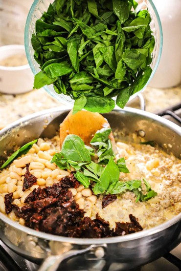 A person transferring fresh spinach leaves into a large silver skillet filled with white beans, sun-dried tomatoes, and sausage in a brown sauce.