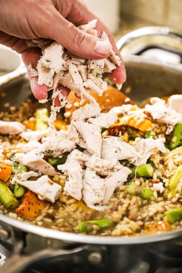A person adding pieces of cooked turkey into a skillet filled with cooked risotto and pieces of roasted butternut squash and asparagus.