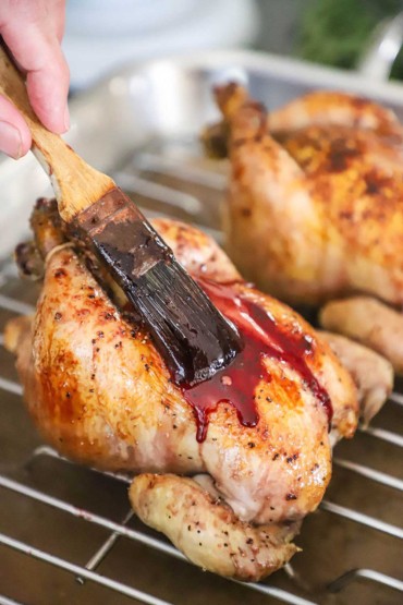 A person using a pastry brush to apply a ruby port glaze to a partially cooked Cornish hen sitting on a baking rack in a roasting pan.