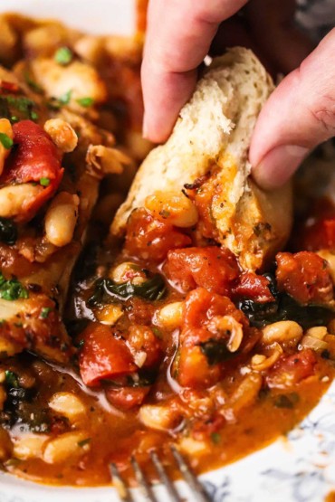 A person using a piece of a loaf of Italian bread to scoop up a Tuscan sauce of tomatoes and beans from a dinner bowl with seared chicken in it.