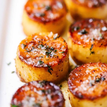 A close-up view of a two rows of fondant potatoes on a white platter and topped with sea salt and fresh herbs.