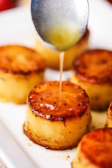 A person using a silver spoon to pour melted butter over a potato sphere that has been seared and roasted.