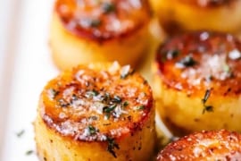 A close-up view of a two rows of fondant potatoes on a white platter and topped with sea salt and fresh herbs.