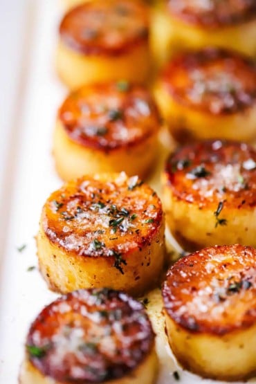 A close-up view of a two rows of fondant potatoes on a white platter and topped with sea salt and fresh herbs.