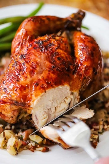 A person using a silver steak knife to cut away a slice of meat from the side of a glazed Cornish hen sitting on bed of wild rice on a white plate.