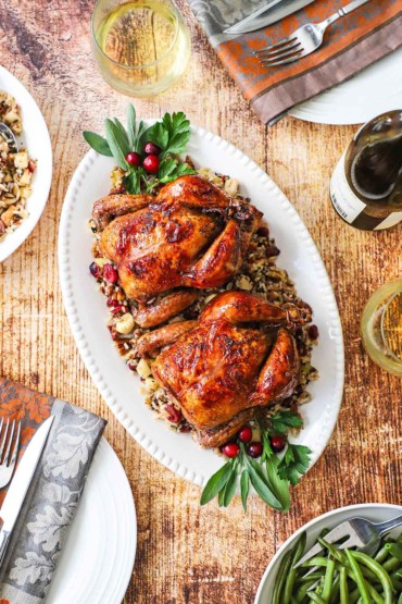 An overhead view of two glazed Cornish hens on a platter with wild rice stuffing flanked by a bowls of green beans and more stuffing.