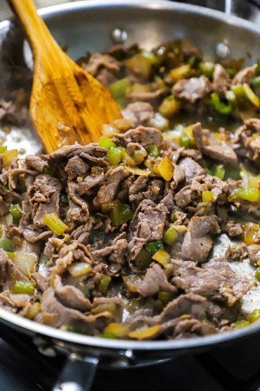 A close-up view of thin strips of ribeye steak and sautéed onion and green bell pepper pieces cooking in a silver skillet.