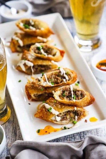 An overhead view of a platter of four Philly cheesesteak egg rolls that have been split in half and drizzled with a sour cream sauce.