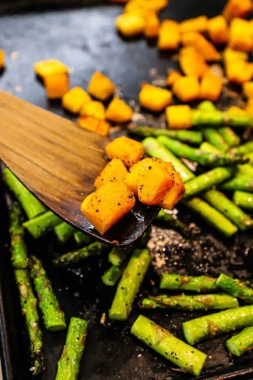 A person using a wooden spatula to lift up several pieces of roasted butternut squash over a baking sheet filled with more of the squash and pieces of roasted asparagus.