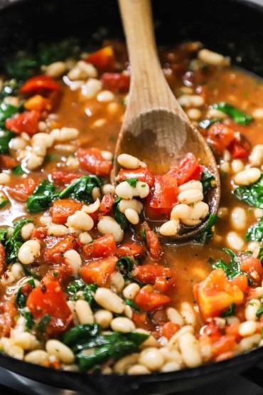 A wooden spoon in a cast-iron skillet that is filled with a simmering sauce of white beans, spinach, and tomatoes.