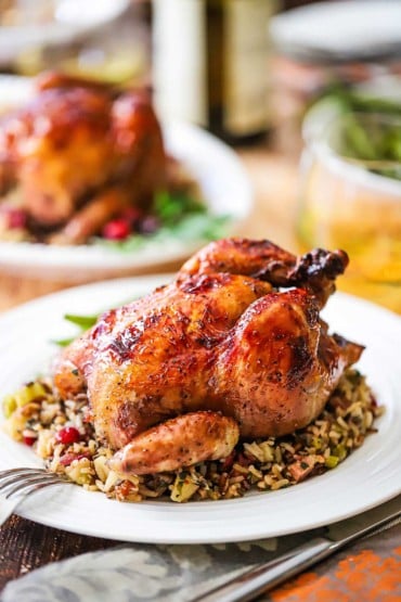 A straight-on view of a glazed Cornish hen sitting on a bed of wild rice pilaf all on a white dinner plate with a fork nearby and another plate of the same in the background.