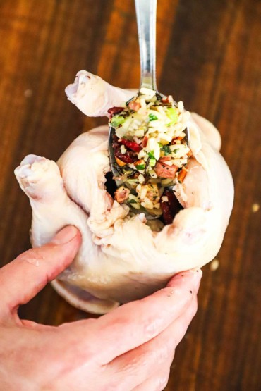 A person holding an uncooked Cornish hen upright while stuffing the cavity with a wild rice with pecans and cranberries stuffing.