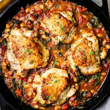 An overhead view of a large black cast-iron skillet filled with four pieces of Tuscan chicken with white beans and spinach.