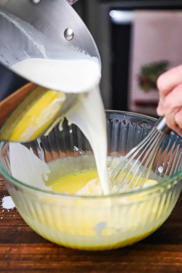 A person pouring warm milk into a bowl of tempered egg yolks that are being whisked.