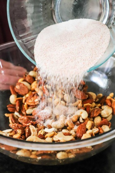 A person pouring a spiced sugar mixture from one large glass bowl into another glass bowl filled with assorted nuts that have been coated in frothy egg whites.