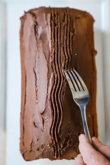 A person using the end of a dinner fork to form bark-like formations in chocolate icing on top of a yule log.