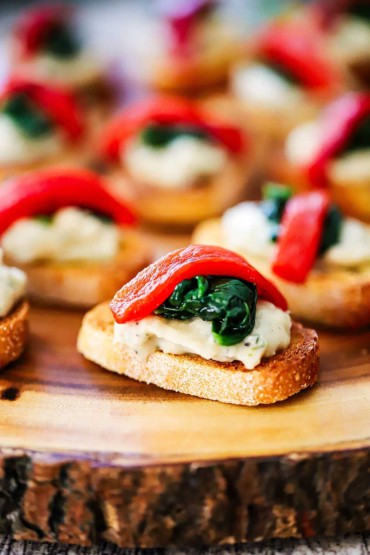 A close-up view of Christmas crostini with a layer of puréed white beans topped with a small amount of sautéed spinach and that topped with a strip of roasted red bell pepper.