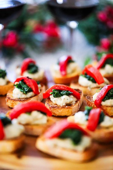 A side view of a platter filled with small toasted crostinis topped with a bean purée, sautéd spinach, and a strip of roasted red bell pepper.