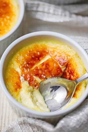 An overhead view of a small white ramekin that is filled with a broken crème brûlée with a spoon inserted into the custard through the crisp sugar top.