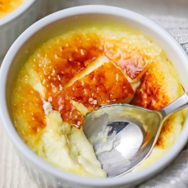 An overhead view of a small white ramekin that is filled with a broken crème brûlée with a spoon inserted into the custard through the crisp sugar top.
