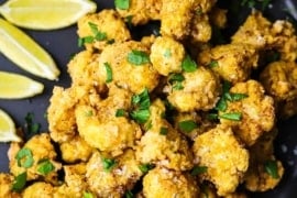 An overhead view of a black circular plate filled with a pile of fried cauliflower with lemon with a row of lemon wedges off to the side of the plate.